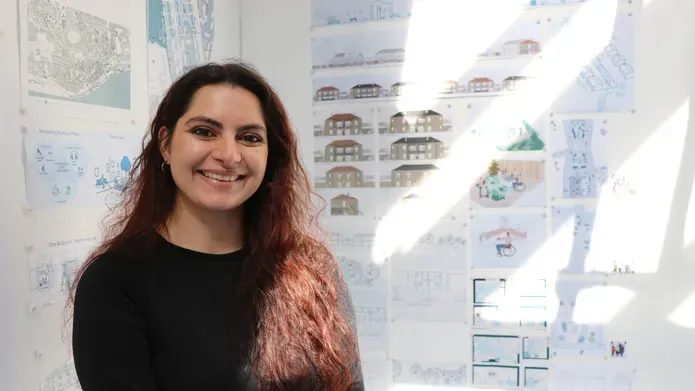 Young woman with long dark hair looking at camera and smiling, with architecture drawings on a wall behind her