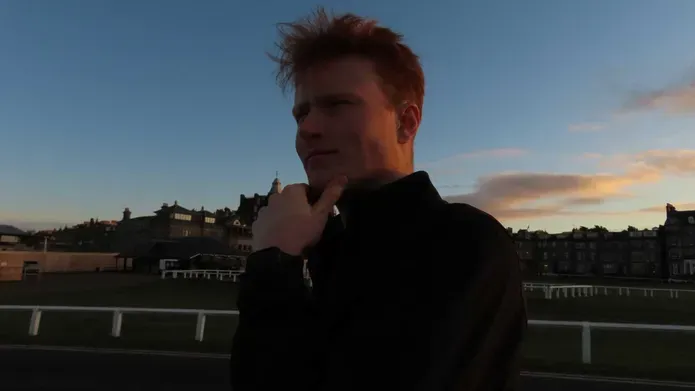 Jarret Fiske with his hand on his chin, looking thoughtful, there is grass behind with white fencing and buildings.