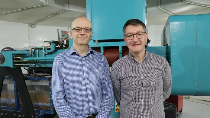 two men wearing checked shirts and glasses, looking and smiling at camera, with large machinery behind them