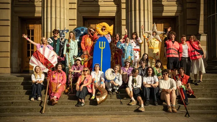 A large, costumed group poses on the steps of Caird Hall in the sunshine, many dressed in colourful handmade outfits.