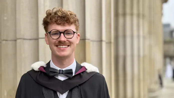 Graduate Kieran, wearing a graduation robe and standing on Caird Hall steps