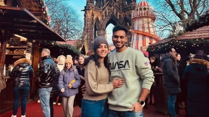 A young couple posing together at a busy Christmas market, male wearing a grey jumper and female wearing a cream jumper with a hat.