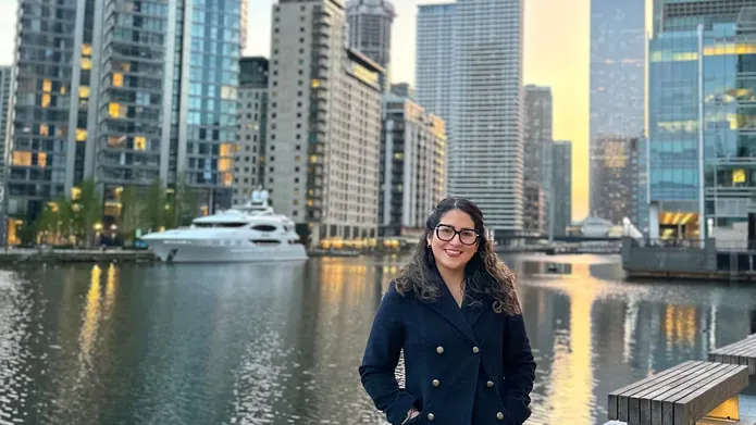 Jeanett stands next to a canal with a yacht in the background and tall high rise buildings on each side of the water
