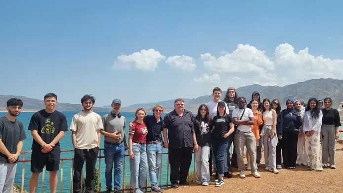 A large group of students, standing in a line smiling at the camera, standing outside with a lake and mountains behind them
