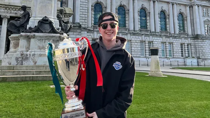 Student wearing a black jacket, grey hoodie, backwards cap, sun glasses and smiling at the camera holding a trophy outside