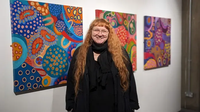 Artist Janice Aikten stands smiling in front of 3 of her colourful canvases