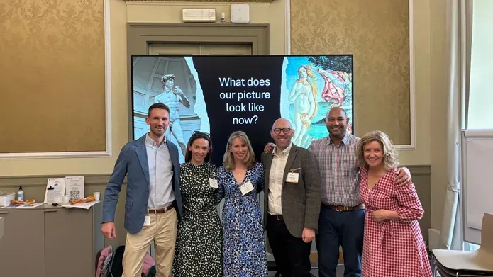 A group of people stand shoulder to shoulder, there is a large screen behind them.