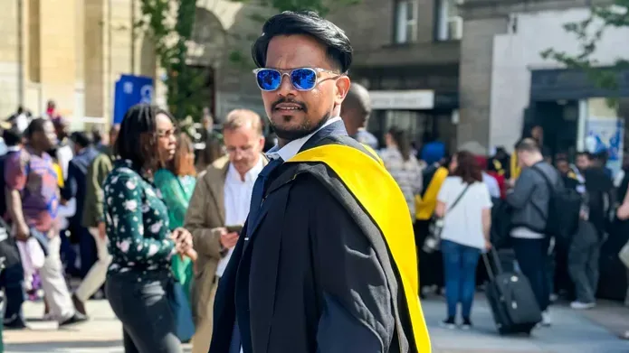 Mohammed Suhail wears sunglasses and graduation robes with a graduation yellow sash in a crowd of people