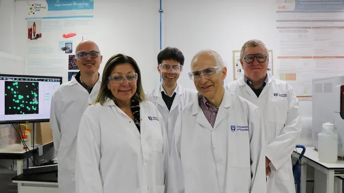 five scientists wearing lab coats and safety glasses, standing next to computer and lab equipment, looking and smiling at the camera