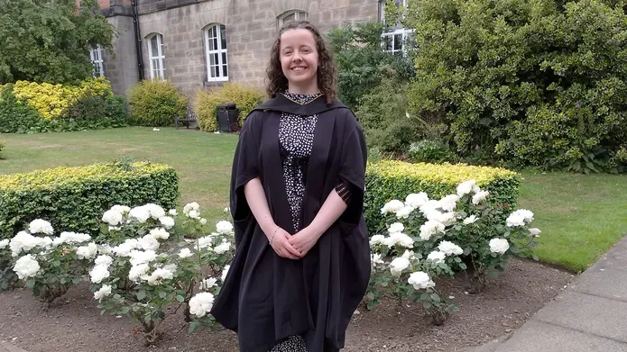 woman standing on garden path with flowers and grass behind her and an older looking house in the background. She is wearing a graduation gown and smiling at the camera
