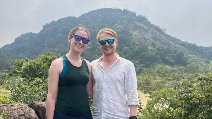Two people wearing sunglasses stand in front of a mountain