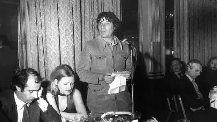 Black and white photography of John Berger, a white middle-aged-man, standing at an awards ceremony, speaking to the audience seated around tables. He holds a piece of paper he is reading from.