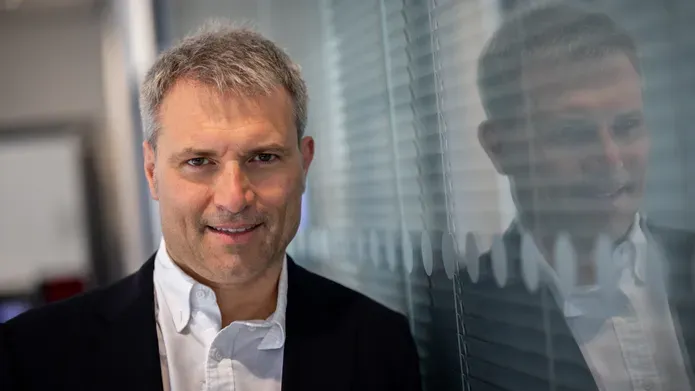 Alessio Ciulli in black jacket, leaning against a glass window, with blinds closed behind it.