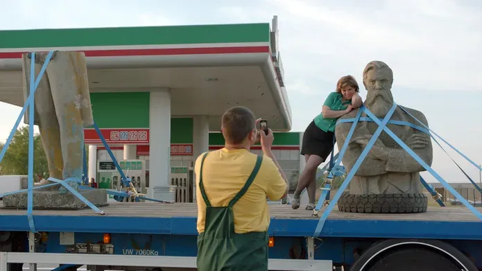 Film still. A sculpture of Frederick Engels is strapped to the back of an open topped truck. It is stopped at a petrol station. A woman leans on the status' shoulders while a man takes a photo of her on the truck leaning on Engels