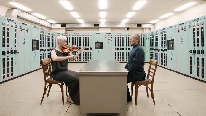 Film still. Two protagonists sit facing each other across a table. One is playing a violin and the other is dressed as a diplomat. They are in an engine control room surrounded by switches and buttons