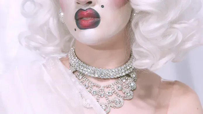 Close crop colour photo of a glamorously made-up person. White face-powder, full-red and black lipstick, beauty spots, sliver necklace and white blond hair