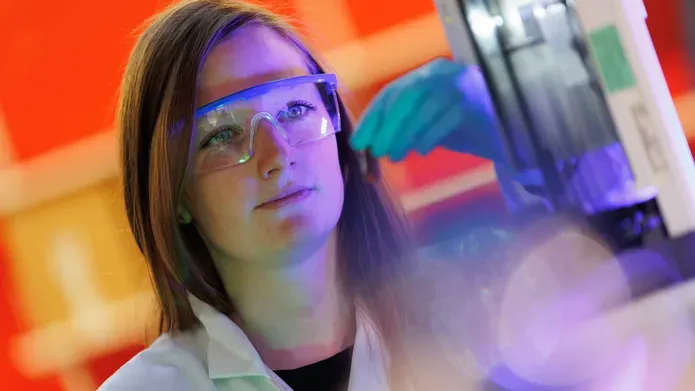 Head and shoulders shot of woman with medium length dark hair, wearing safety glasses and a lab coat, looking at a computer screen