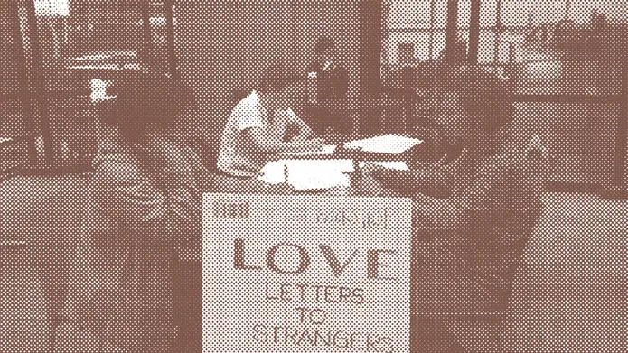 Sepia toned photograph of workshop participants at desks writing. A sign says Love Letters to Strangers