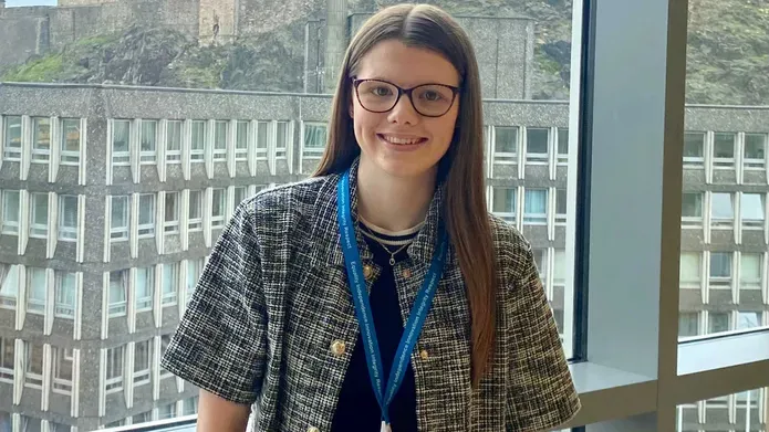 Student wearing glasses, infront of a window and smiling at the camera, wearing a chequered jacket and a black top.