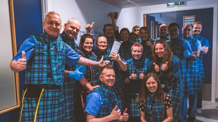 A group of people wearing blue and tartan scrubs stand in a corridor with white walls and blue doors.