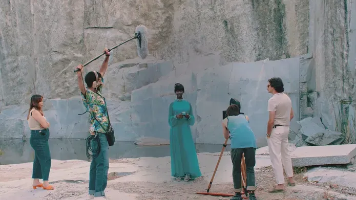 Film still. A film crew circle a Black woman in a long blue dress. They are shooting outdoors in a quarry