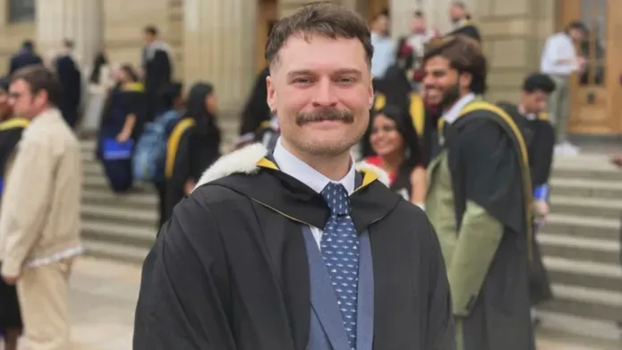 student wearing a blue suit with a graduation gown smiling at the camera