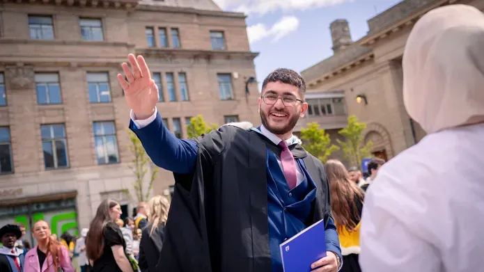 A graduate in graduation dress standing in the City  Square waving to someone 
