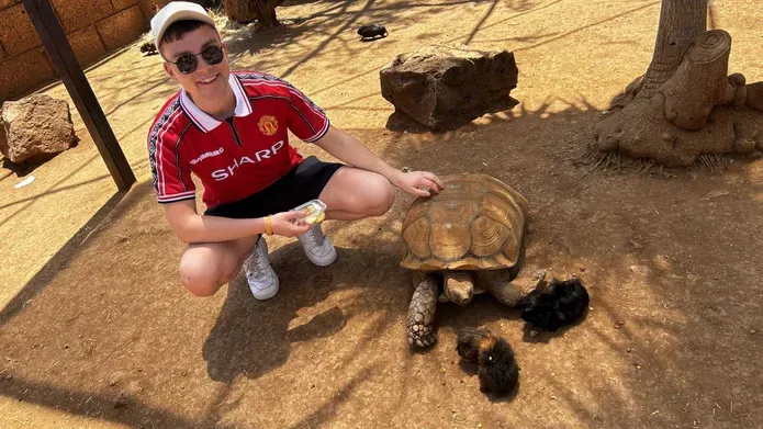 Aidan Harper ewars a red football top, shorts and a cap with sunglasses. He squats down to pet a tortoise, there are also guinea pigs.