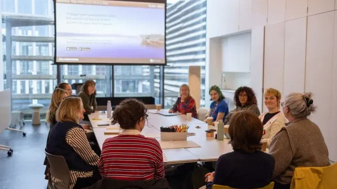 A group of people sitting around a large table with a view of the V&A outside the window 