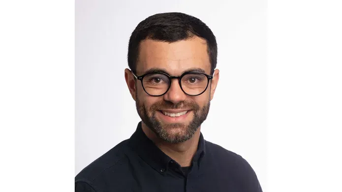 A head and shoulders photo of a man with short brown hair, beard and glasses smiling