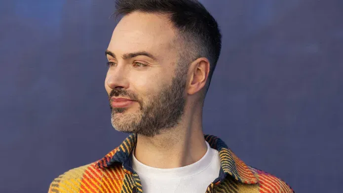 A man with dark hair and facial hair, stands in front of a blue wall, looking to the side.