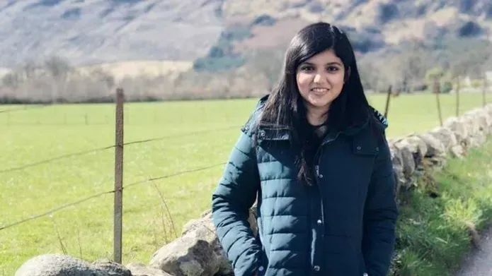 A student smiling and posing for the camera, with a jacket on with fields and hills in the background.