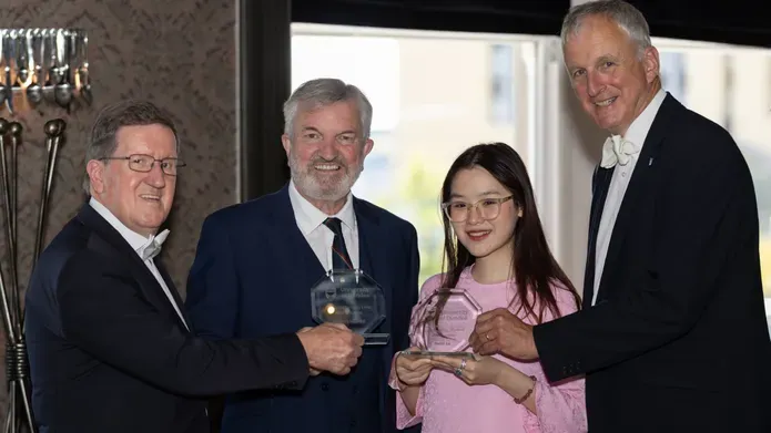 Four people smiling and holding clear glass Bridge Alumni Awards. From left to right: George Robertson, John Fyffe MBE, Heidi Lee, and Nigel Seaton. The group stands indoors with soft lighting and patterned wallpaper in the background.