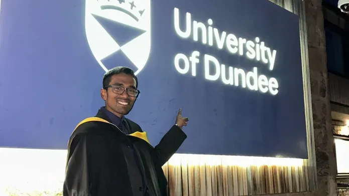 Graduate in his graduation gown pointing at the University of Dundee sign which is lit up in the evening.