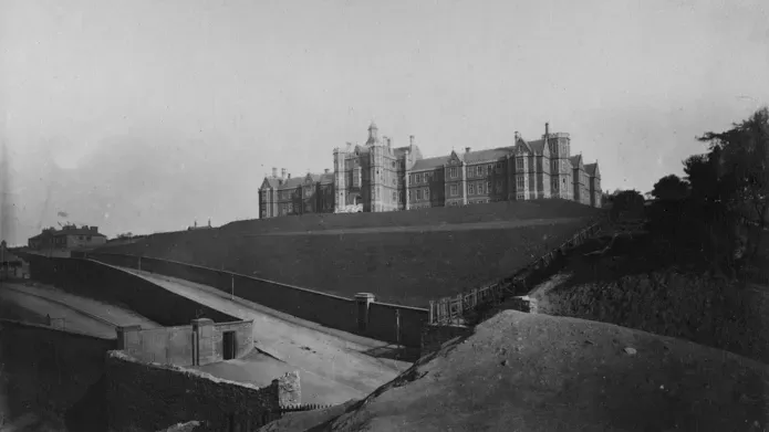 A distant view of a large stone hospital building on a hill, with sloping roads and stone walls in the foreground.