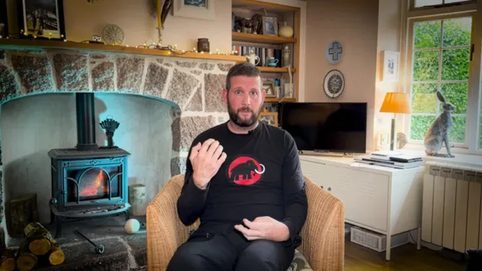 man sitting on sofa, holding hands up, in living room with wood burning fire behind