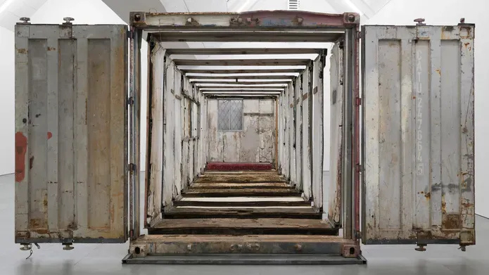 Exhibition view. Tight crop showing the end of a large sculpture. A shipping container cut into sections and laided out in an expanded view