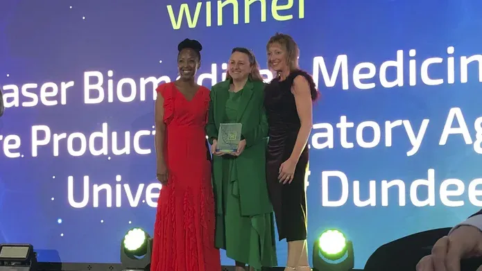 Three women stand on a stage, a large screen is projected behind them with the word "winner" directly above their heads.