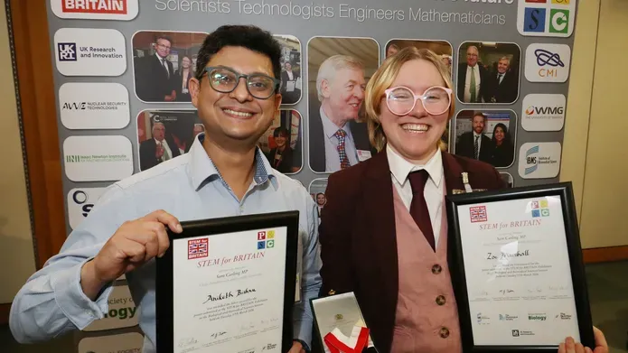 Two people stood smiling holding framed certificates stood in front of a pop up banner. One individual is also holding a medal in a box.