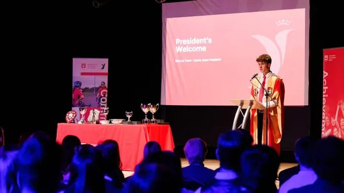 A young man in academic robes gives a speech at a podium, with trophies on display and an audience watching