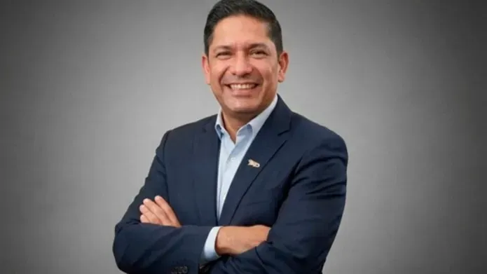 Juan Carlos Hurtado Parra wearing a dark blue suit with a pale blue shirt, he has his arms crossed and he is smiling.