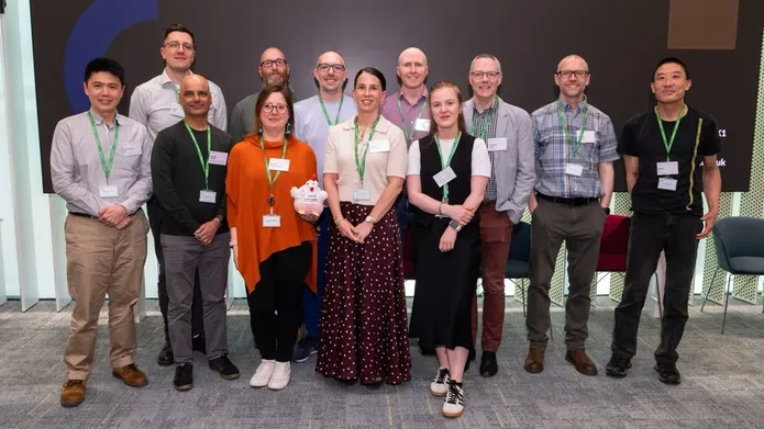 Group photo of researchers and staff wearing name badges, standing together at a conference or showcase event, with presentation screens in background