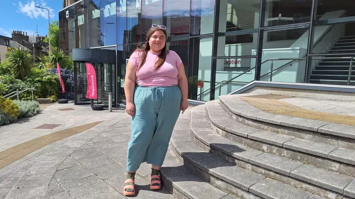 woman wearing pink top and green trousers, standing on steps outside of a glass bulding