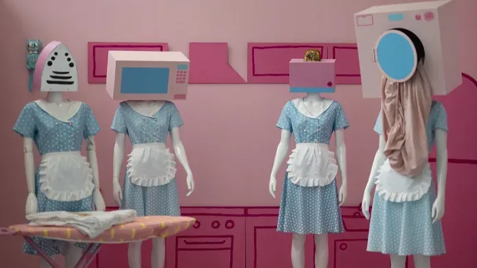 Light pink walls and carpet with darker pink cartoon style furniture made from cardboard and stuck to the walls. Four mannequins in front wear blue 1950s style dresses and white aprons, with kitchen equipment for heads 