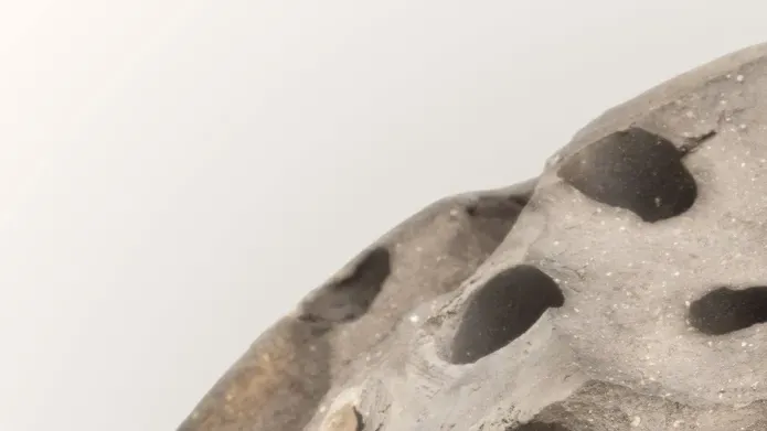 A section of a grey concrete looking sphere, with large holes, against a pale grey background.