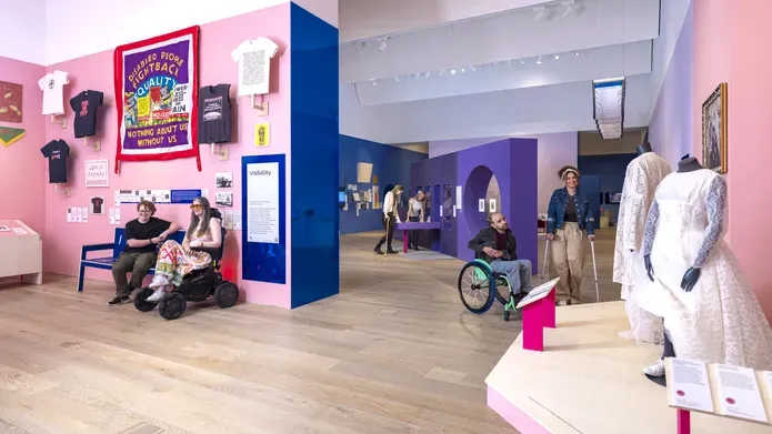 A iew of the exhibiton, with pink and purple-coloured rooms. Viewing the exhibits are people in wheelchairs and walking on crutches.