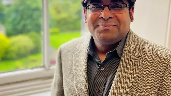 Dr Bannerjee, wearing a blazer and cap, standing next to a window with grass outside.