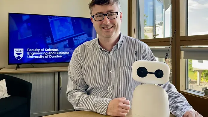 Dr Mike Crabb sitting at a table with a small, white robot, with black camera eyes and two black antenna. In the background is a television screen showing a blue backgrond with University of Dundee branding.