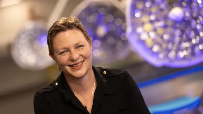 Professor Lucina Hackman in black coloured jacket, smiling. Behind her are three large, circular lights, which are tinged with purple.