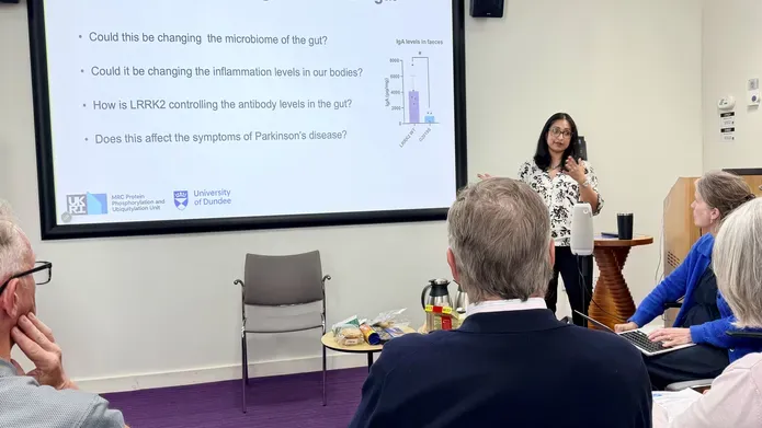 A woman stands at a large smart board, presenting to people sitting down in a room.